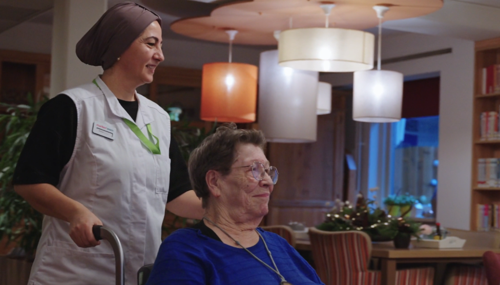 Op deze afbeelding zie je een zorgverlener (vrouw) met een hoofddoek; zij duwt een vrouw op leeftijd in een rolstoel en ze glimlachen allebei.